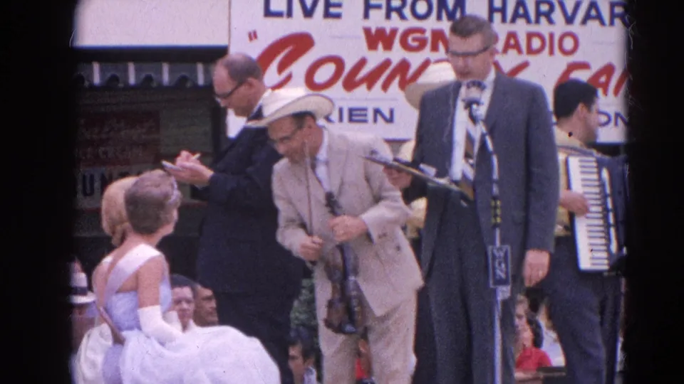 The fair stage shows a violinist, suited announcer, accordion player, and a young dancer near the microphones in Harvard, Illinois, 1964.