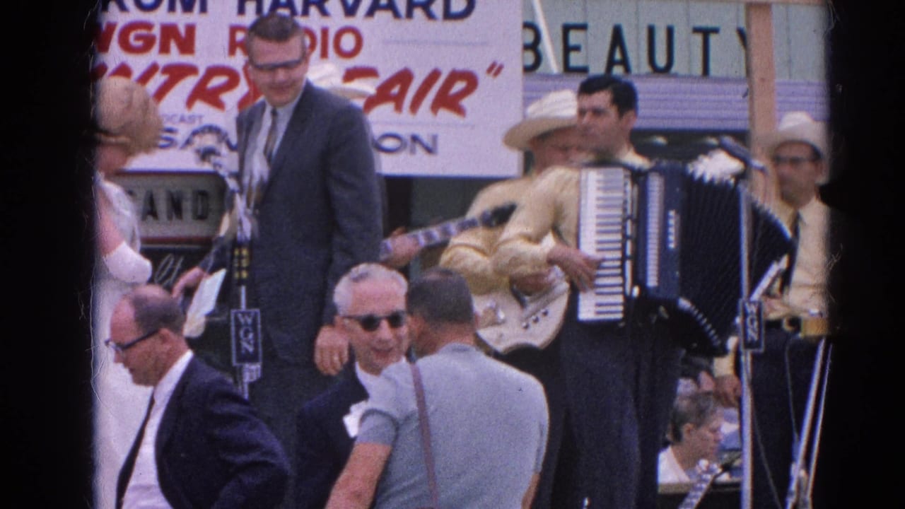 Male host stands at microphones beside musicians and spectators under a country fair banner.