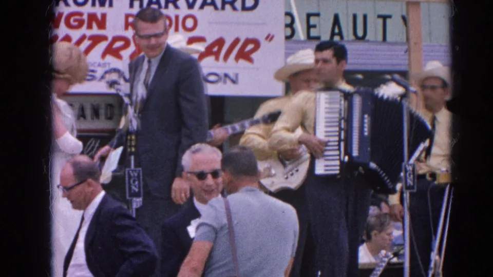 At a 1964 Harvard, Illinois country fair performance, a suited male host stands near microphones while accordion and guitar players perform behind him.