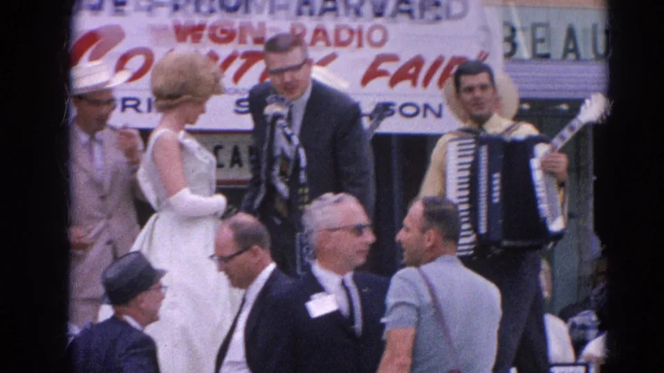 A female singer in a white dress and gloves stands by the suited host as musicians play during the Harvard, Illinois country fair performance.