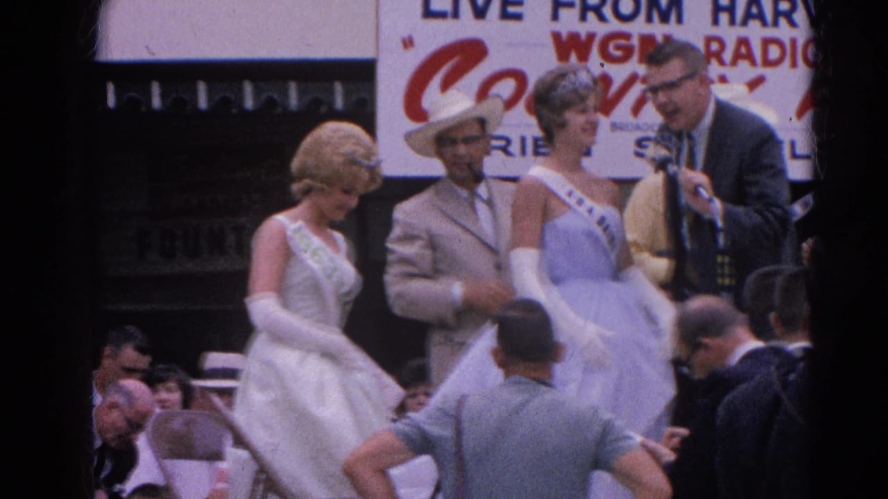 Two sash-wearing women in formal gowns stand near microphones during a public fair presentation.
