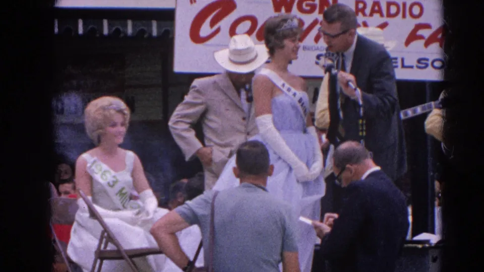 The frame shows a Harvard, Illinois fair pageant presentation with a seated sash-wearing participant, a standing contestant, and radio signage overhead.