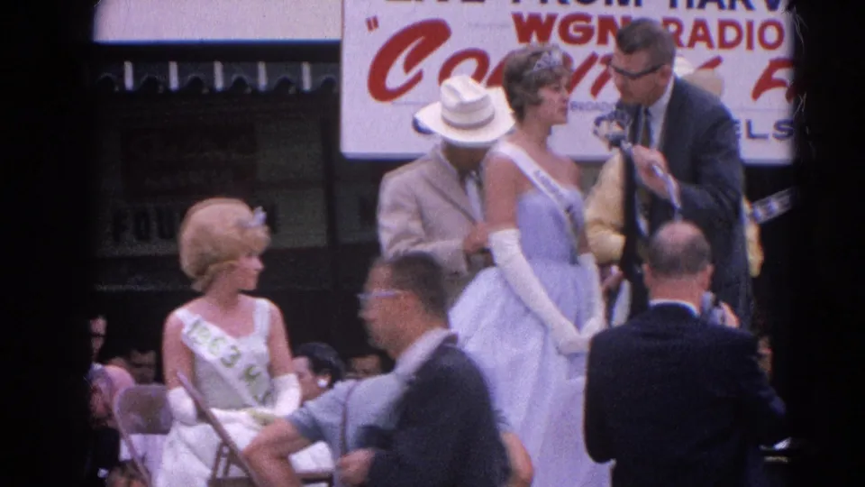 During the 1964 Harvard, Illinois fair pageant interview, a passerby crosses the foreground while contestants and the interviewer remain on stage.