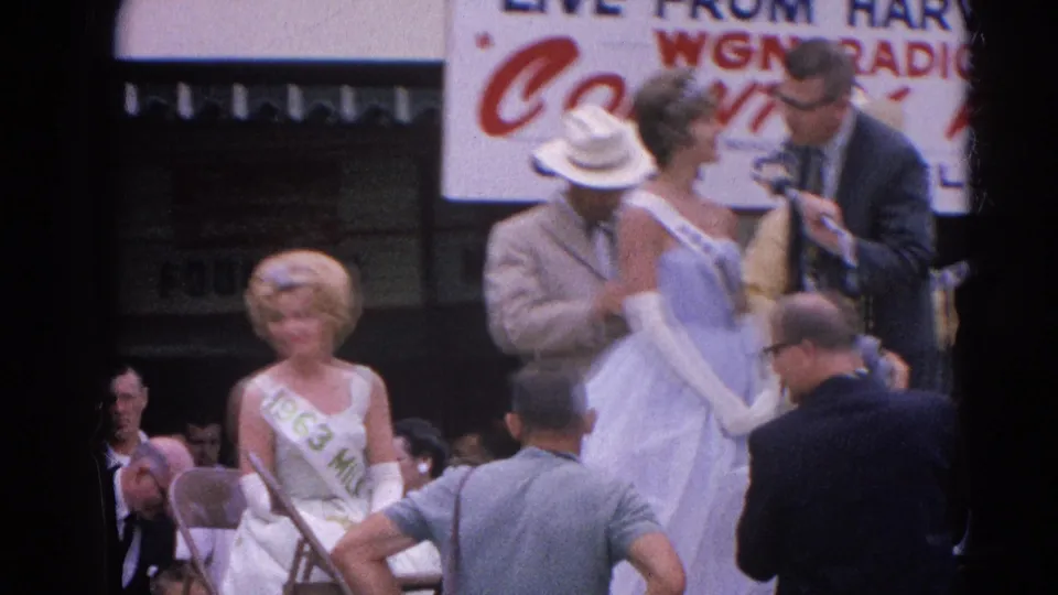 The Harvard, Illinois 1964 fair stage remains crowded as the blue-gowned contestant, interviewer, seated sash contestant, and bystanders fill the frame.