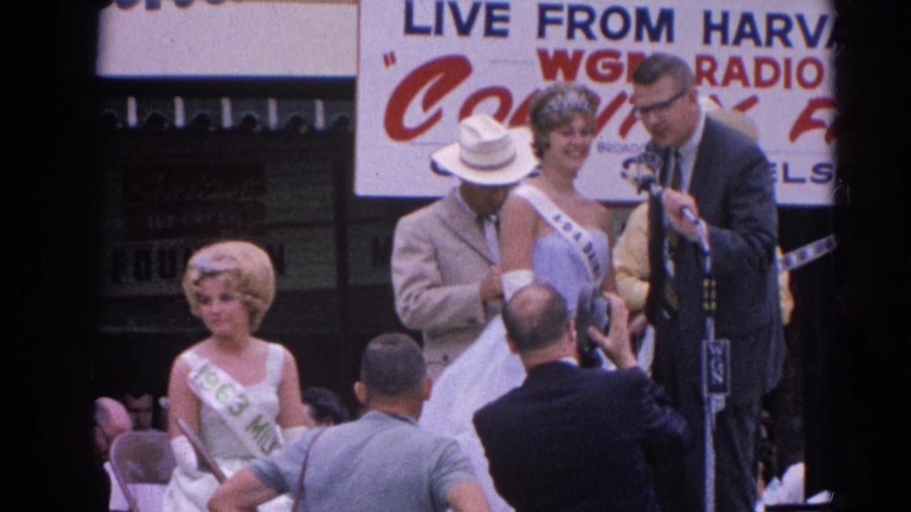 Announcer at microphones beside pageant contestants on a fair stage with photographers in front.