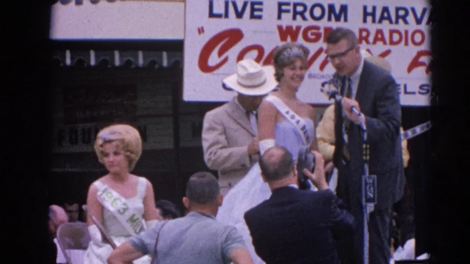At the Harvard, Illinois fair stage, a suited announcer stands by microphones as pageant contestants in white gowns and sashes face the crowd.