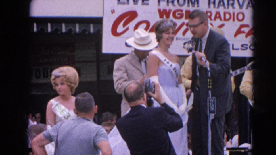 A suited man speaks at a stage microphone while two sash-wearing pageant contestants stand nearby at the 1964 Harvard, Illinois fair.