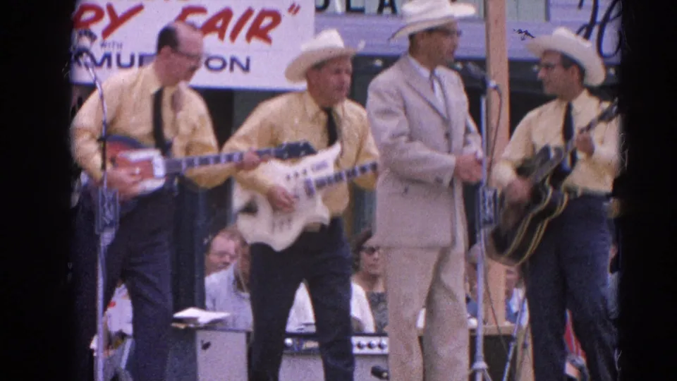 Musicians in matching yellow shirts and cowboy hats play electric guitars on the fair stage, with signage and spectators behind them.