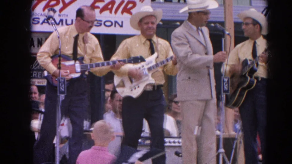 The fair stage shows guitar players in cowboy hats and matching shirts, with a light-suited man standing among them during the performance.