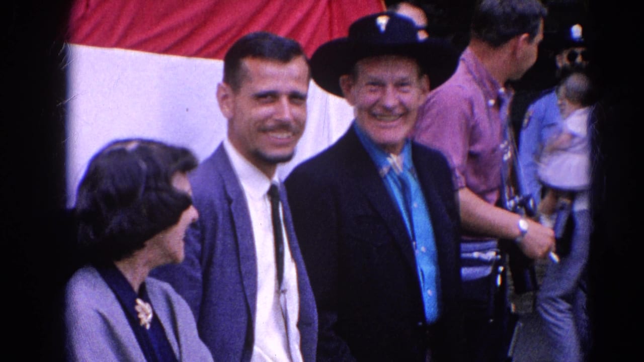 Two men and a seated woman near a large red and white backdrop at an outdoor event.