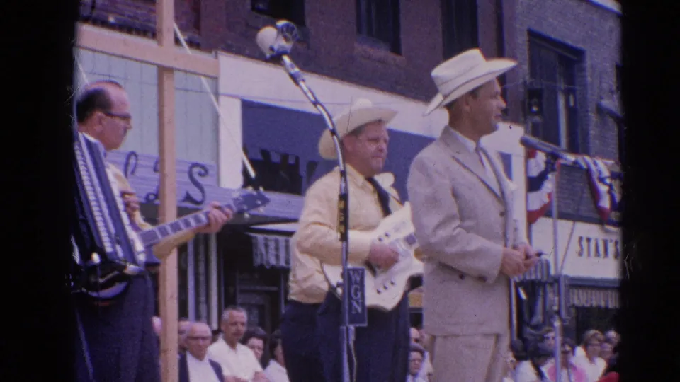 The Harvard, Illinois band performance continues with accordion, electric guitar, cowboy hats, storefront awnings, and spectators visible behind the stage area.