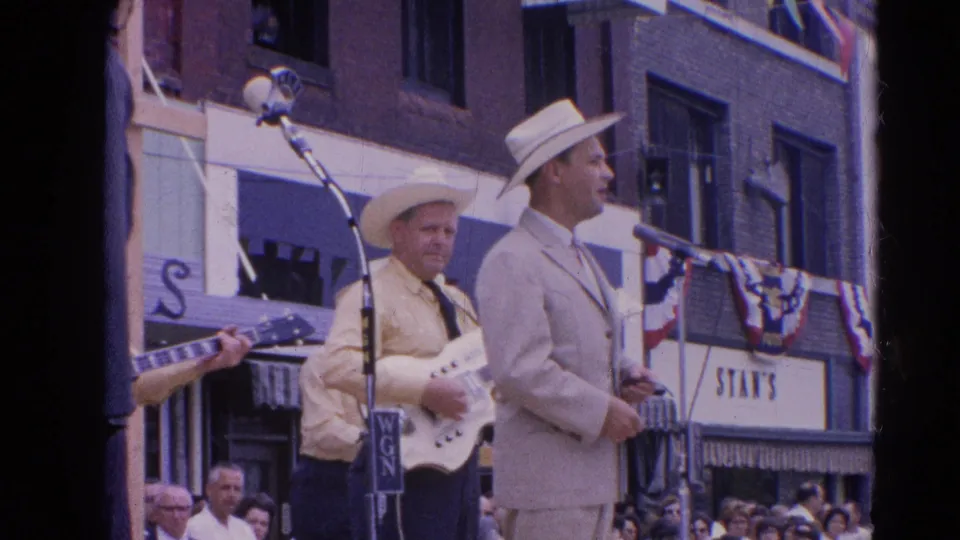 A light-suited singer in a cowboy hat stands by a microphone as a guitarist and crowd appear along a decorated Harvard, Illinois street.