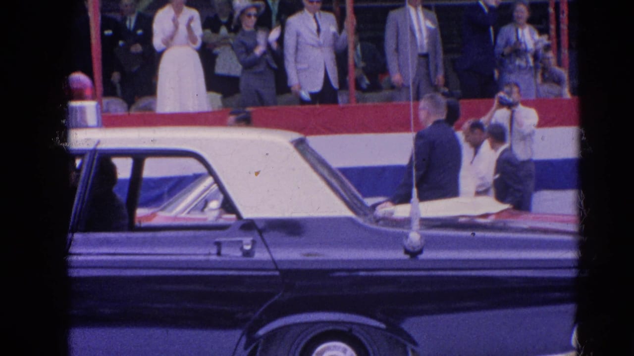 Dark car in foreground with spectators standing behind parade bunting.