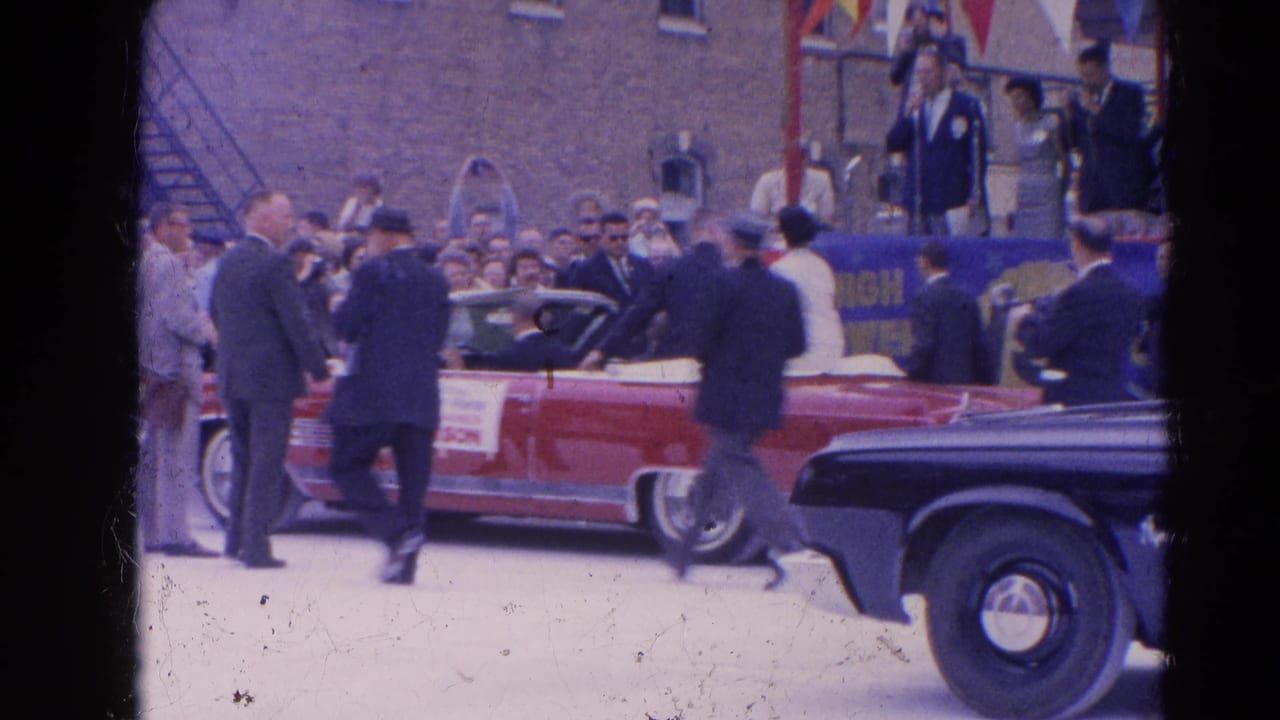 Red convertible surrounded by suited men and spectators during a parade.