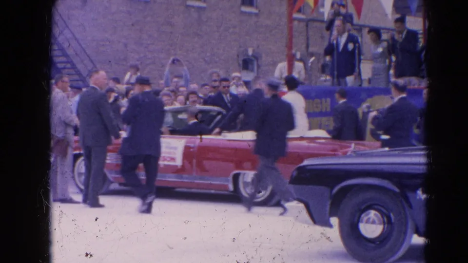 Red convertible with a white top moves through a Harvard, Illinois crowd in 1964 as suited men and spectators gather nearby.
