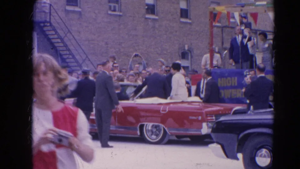1964 Harvard, Illinois parade frame showing a red convertible, a crowd at the curb, and a woman in red and white holding a camera.