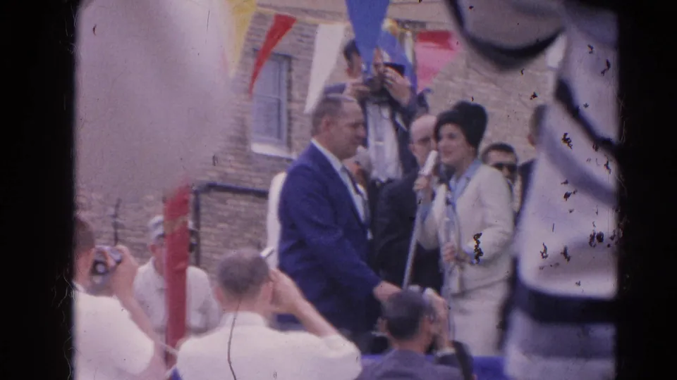 In Harvard, Illinois in 1964, spectators and photographers face a woman in a pale suit standing at microphones beneath bunting.