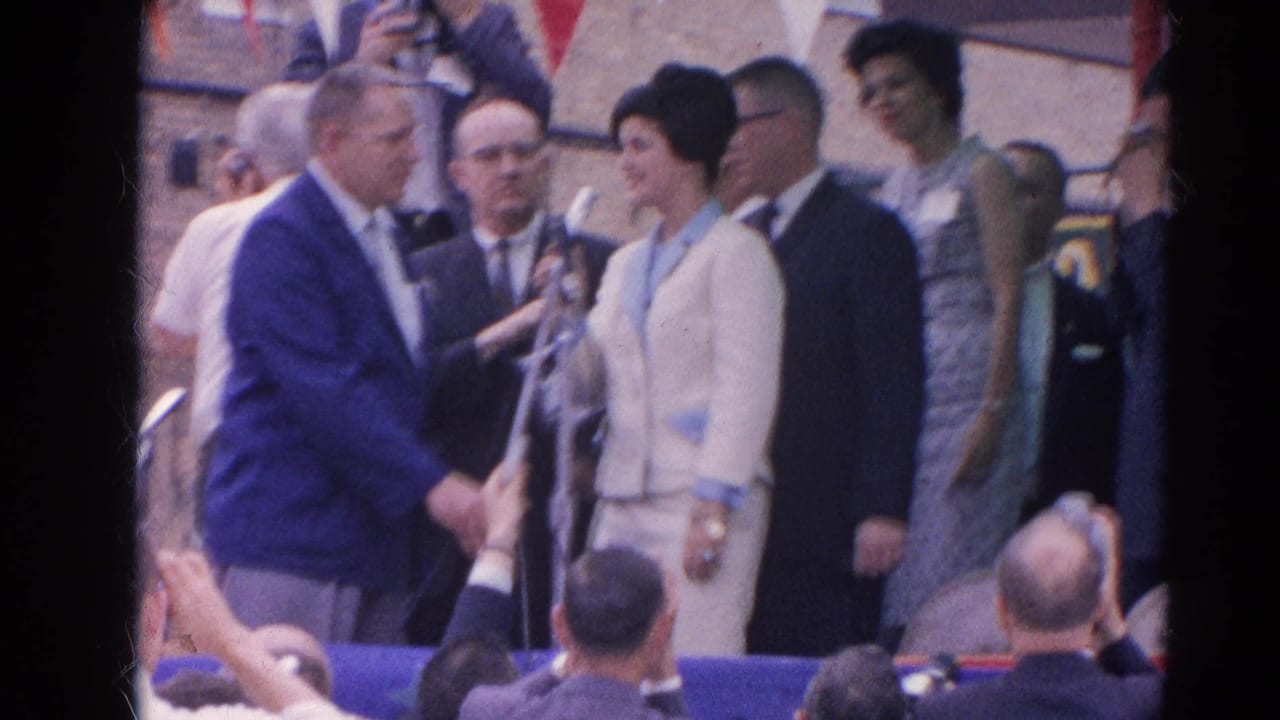 Woman in a light suit speaks at a microphone beside suited men on a decorated outdoor stage.