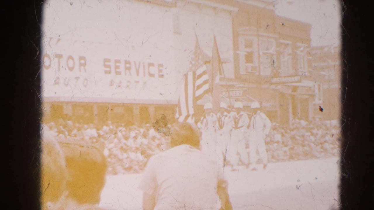 A faded 8mm view of the Harvard, Illinois parade shows a flag bearer before a crowd and storefronts with patriotic bunting.