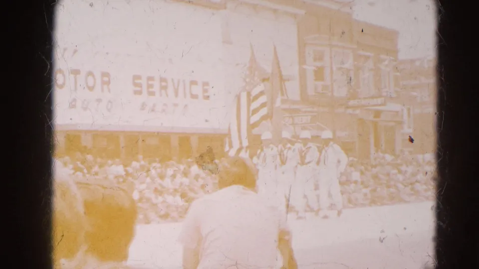 A faded 8mm view of the Harvard, Illinois parade shows a flag bearer before a crowd and storefronts with patriotic bunting.