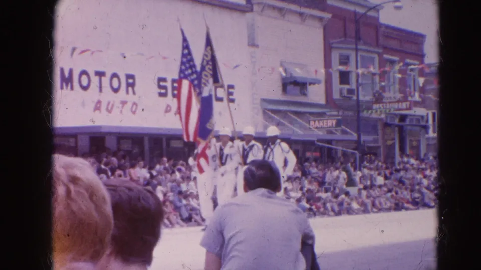 Uniformed marchers in Harvard, Illinois carry American and unit flags past spectators, with Motor Service and Bakery signs visible behind them.
