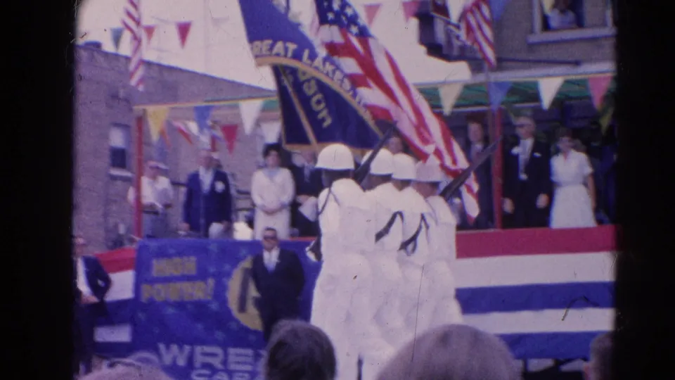 White-uniformed marchers carry American and unit flags past a decorated stage in the 1964 Harvard, Illinois public parade scene.