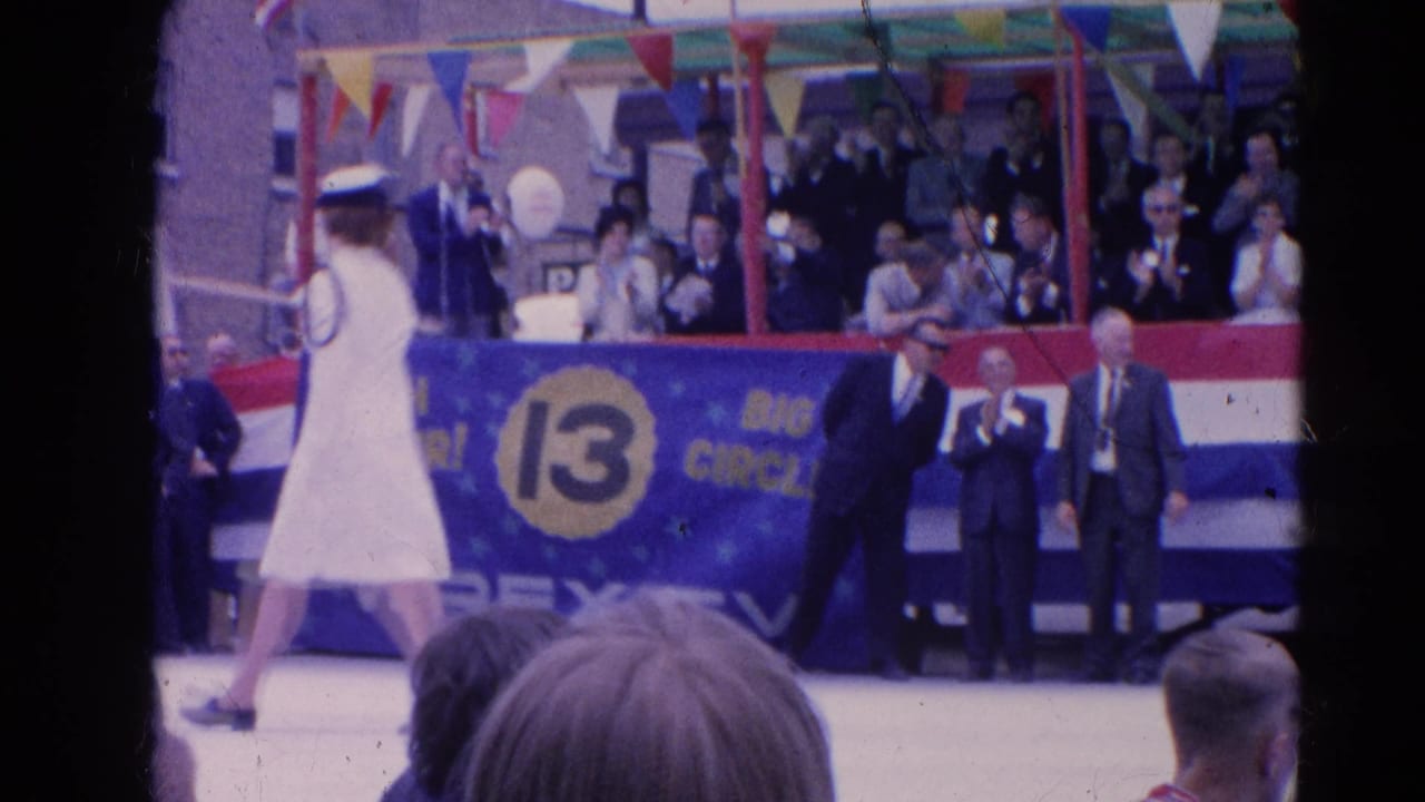 Woman in a white parade uniform walks past a decorated reviewing stand with spectators behind her.