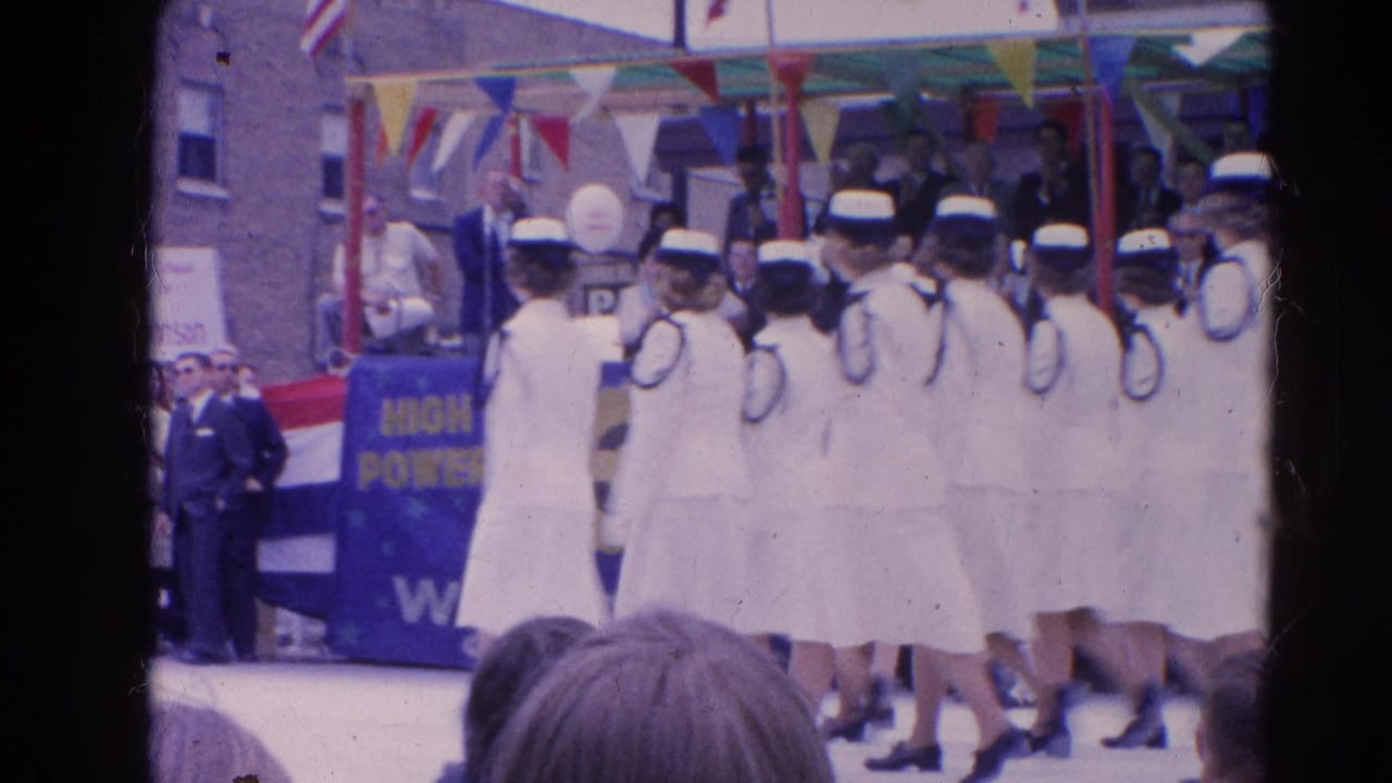 Line of young women in white sailor-style uniforms marches past a decorated stand.