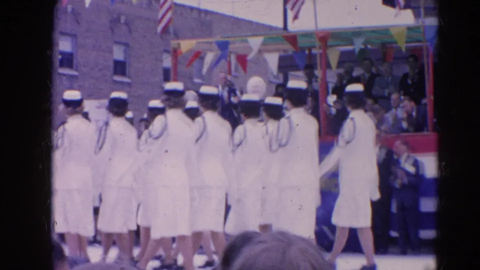 The uniformed marching group continues along the parade route beside a reviewing stand and spectators in 1960s street attire.