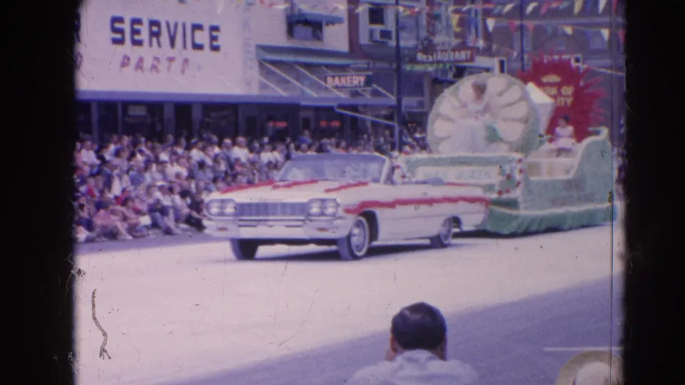A 1960s convertible proceeds through the Harvard, Illinois parade beside a floral float, storefront signs, and a dense sidewalk crowd.