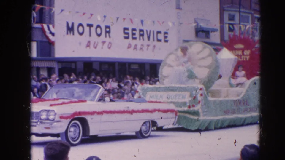 Close 8mm view of the Harvard, Illinois Milk Queen float, with a woman seated above the sign and spectators lining the street.