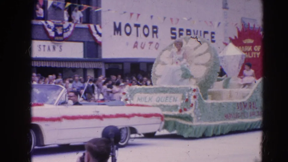 Harvard, Illinois parade frame with the Milk Queen float close to camera, partial convertible, crowd, storefront signage, and visible film scratches.
