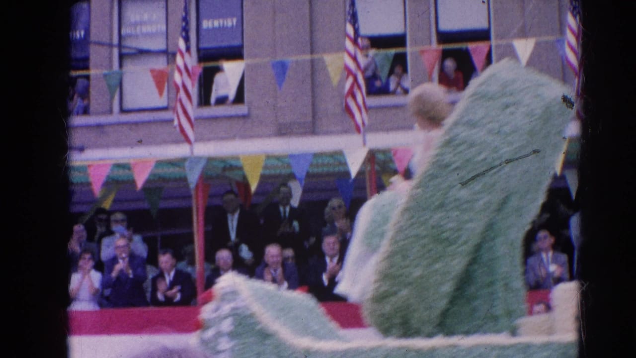 Green parade float passing spectators beneath bunting and American flags.