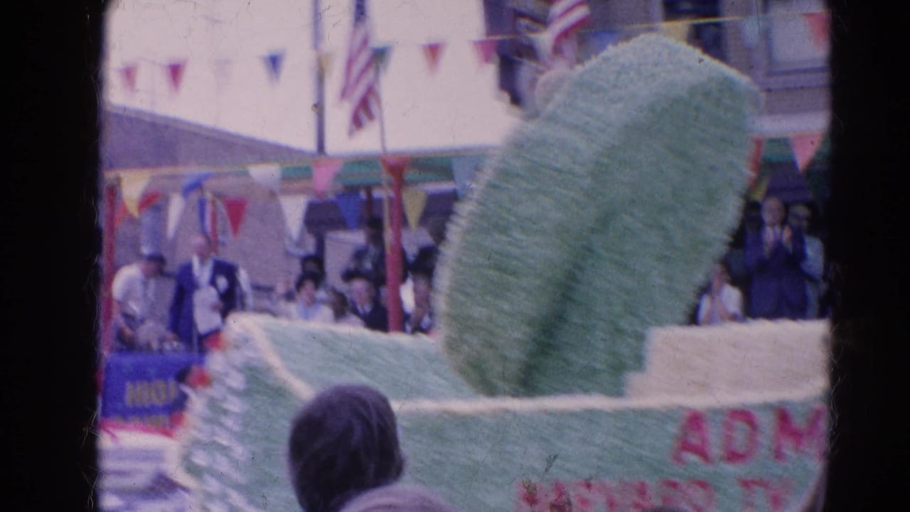 Green parade float with partial text passes through the Harvard, Illinois street scene as spectators watch from sidewalks and upper windows.