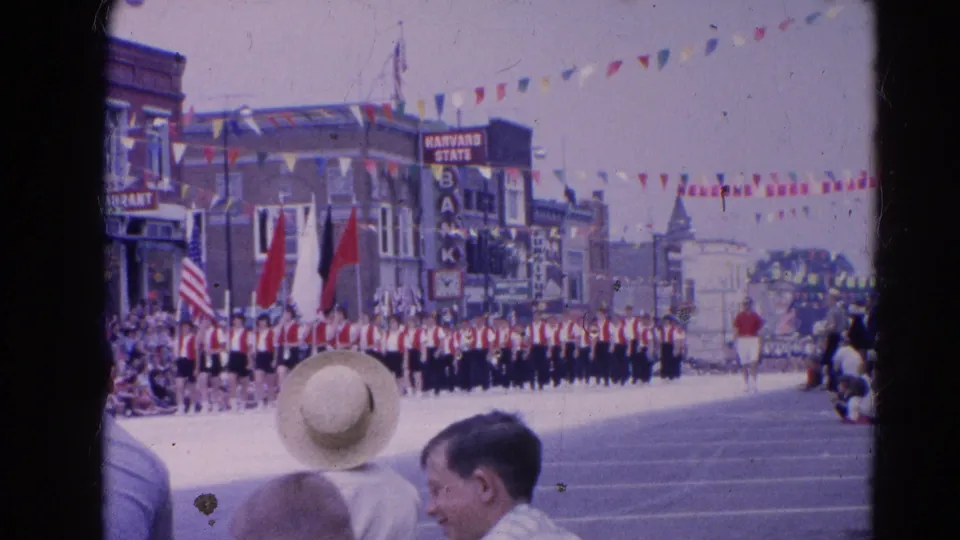 A red-and-white marching band appears down the street during the 1964 Harvard, Illinois parade, with spectators and storefront signs nearby.