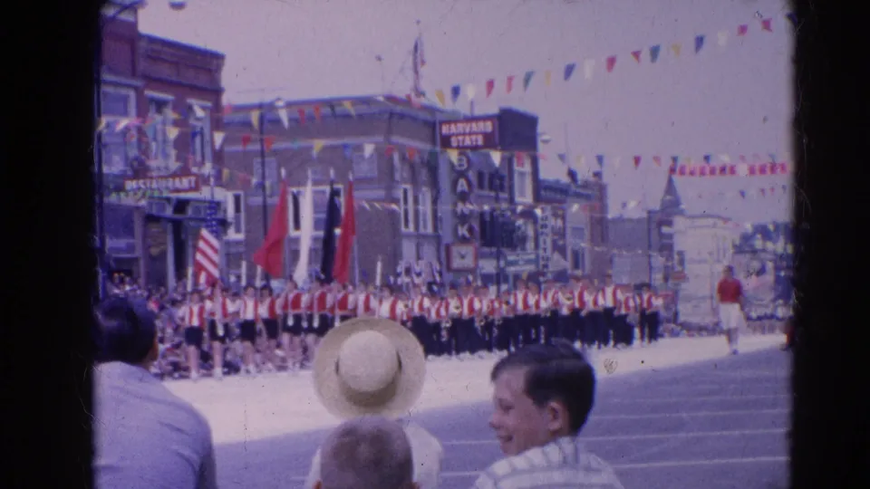 Spectators watch a red-and-white marching band in the Harvard, Illinois parade, with bunting, flags, and downtown storefronts visible.