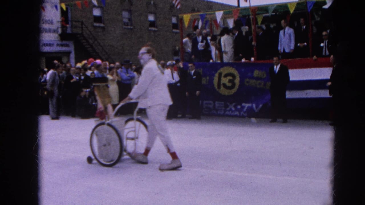 A clown in a white costume pushes a small cart past spectators during a street parade.