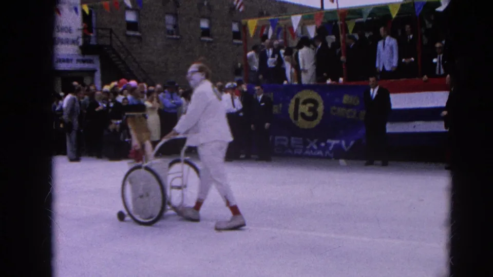 In Harvard, Illinois in 1964, a white-costumed clown pushes a small cart along a parade route in front of bunting, signs, and spectators.