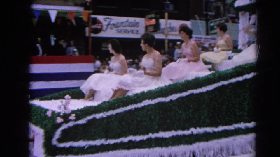 Women in white and pale pink formal dresses ride a decorated float in the 1964 Harvard, Illinois parade, with spectators and signage behind them.