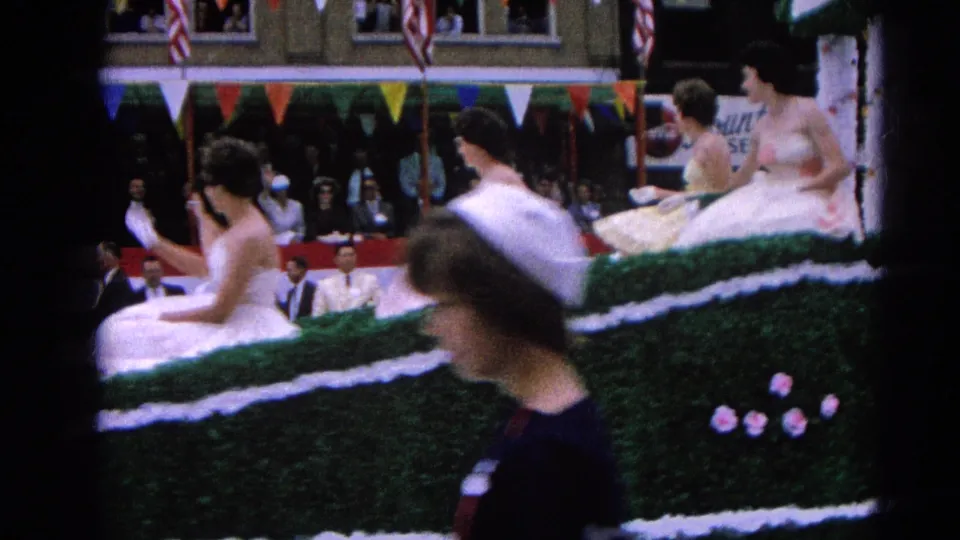 The Harvard, Illinois parade float continues past spectators, showing seated women in formal dresses and a foreground passerby in a dark outfit and white hat.