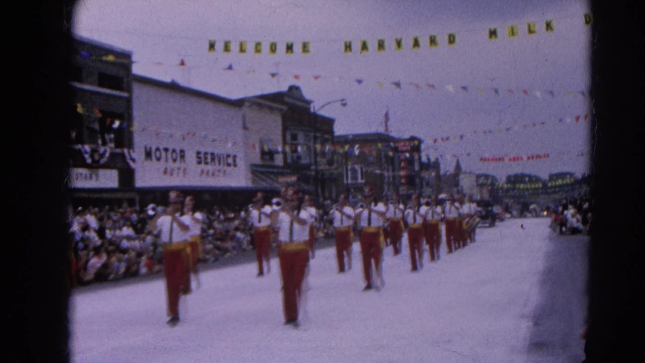 Marching band advances down a parade street beneath a Welcome Harvard Milk Day banner.