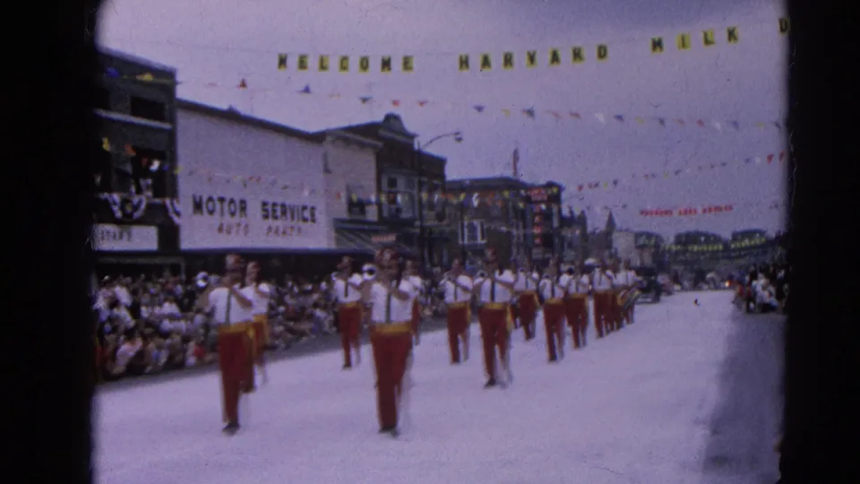 1964 Harvard, Illinois parade view with a marching band approaching under a Welcome Harvard Milk Day banner and spectators on both sidewalks.