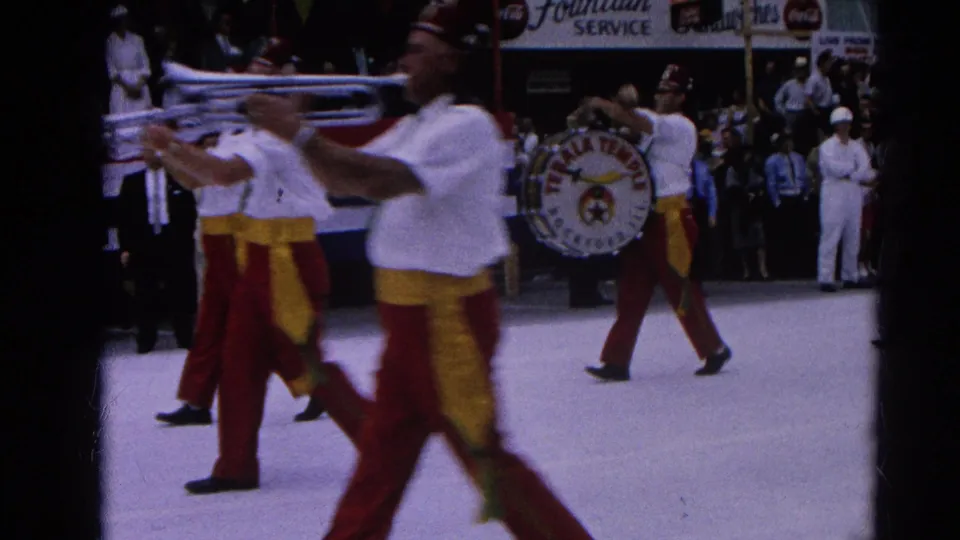 Close Super 8 view of marching musicians passing the camera, including a visible Tebala Temple Rockford, Ill. drum in the parade line.
