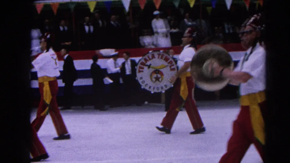 Parade band members pass close to the camera in Harvard, Illinois, with a cymbal player and Tebala Temple drum visible near the crowd.