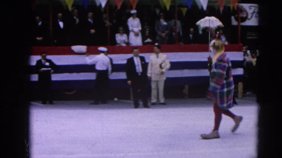 A pedestrian in a colorful plaid coat and hat crosses the foreground as spectators watch the 1964 Harvard, Illinois parade.