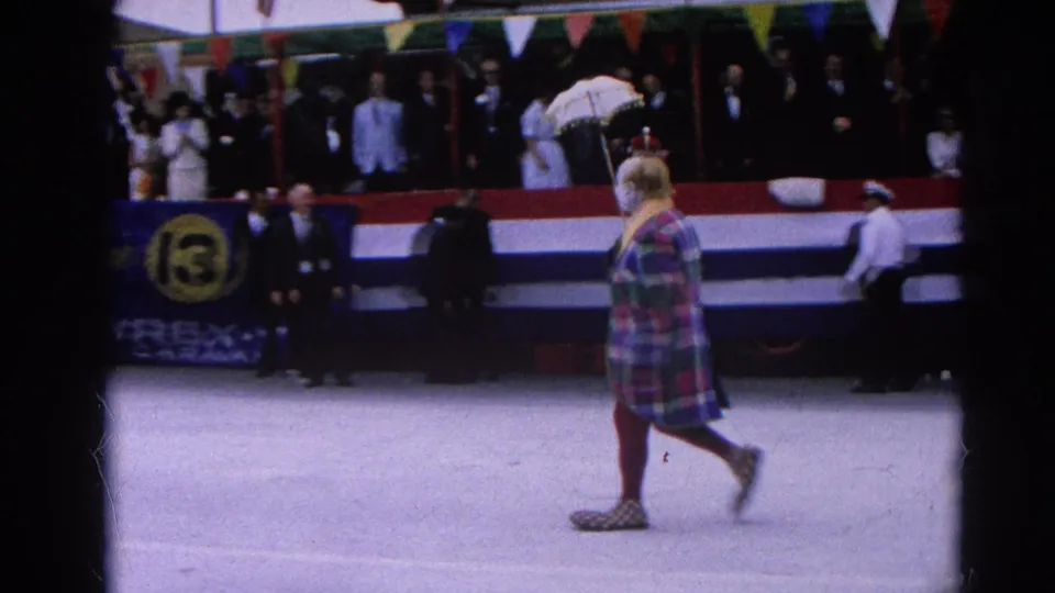 The plaid-coated pedestrian continues across the parade scene with spectators, bunting, and pennant flags visible behind them.