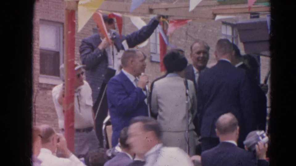 1964 Harvard, Illinois home-movie frame showing a suited man speaking at a microphone during an outdoor civic gathering.