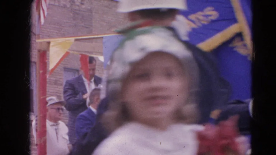 A ceremonial flag bearer in a white helmet crosses the foreground of the 1964 Harvard, Illinois gathering, with spectators behind.