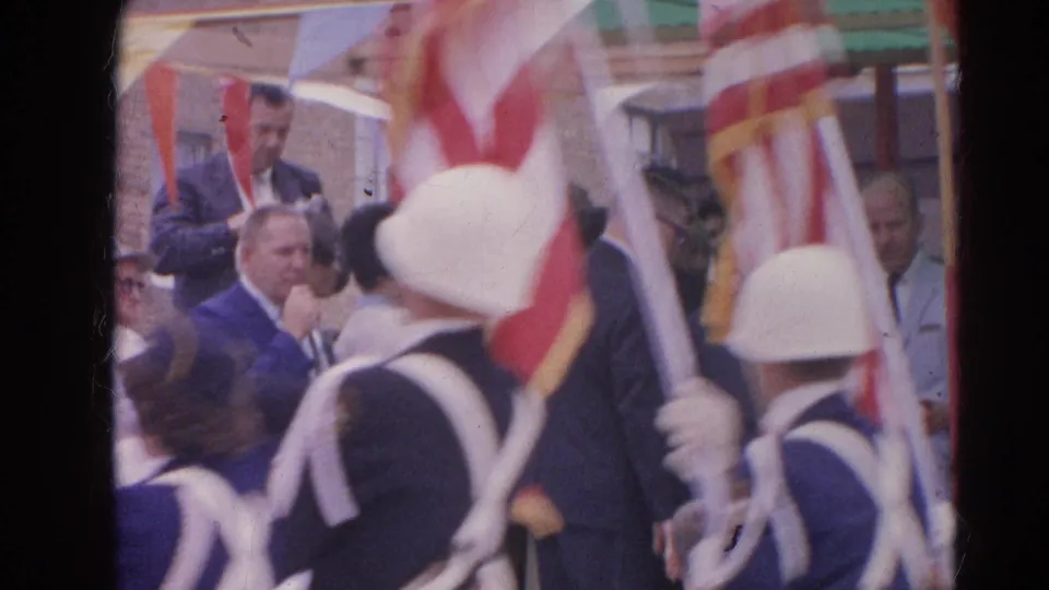 Flag bearers in white helmets move through the foreground during the Harvard, Illinois outdoor civic event, with bunting and suited attendees behind.