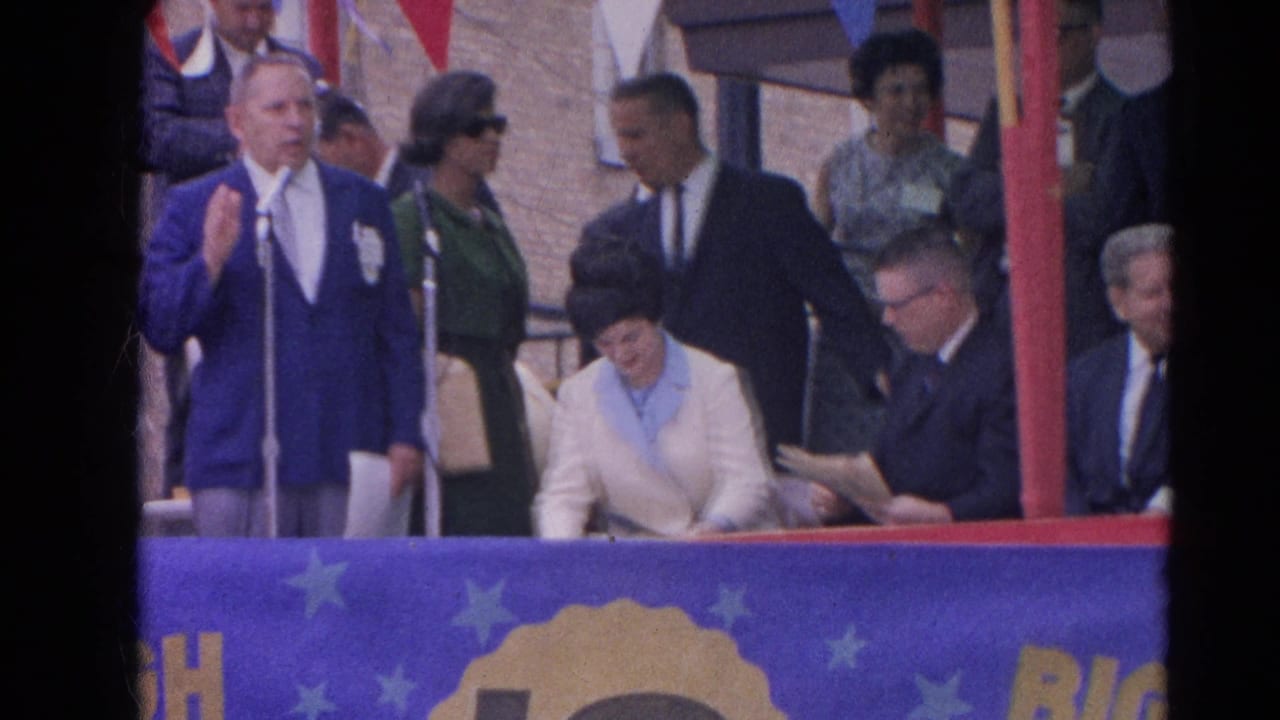 Man in a blue suit speaks at a microphone beside seated and standing attendees on a decorated platform.